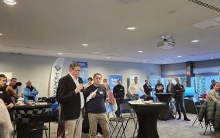 A speaker at the microphone addressing an audience in a large reception hall during the event.