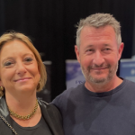 A medium shot of Christelle Marsot and Manu Martinez smiling together at the Techno Piscine 2025 event, with event banners and a trade show background.
