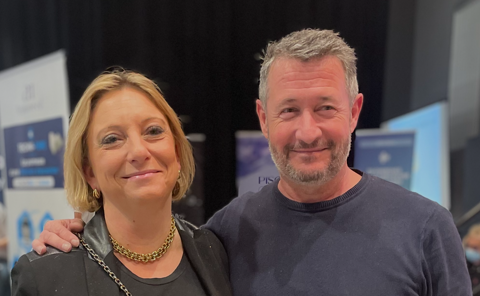 A medium shot of Christelle Marsot and Manu Martinez smiling together at the Techno Piscine 2025 event, with event banners and a trade show background.
