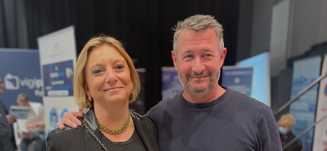 A medium shot of Christelle Marsot and Manu Martinez smiling together at the Techno Piscine 2025 event, with event banners and a trade show background.