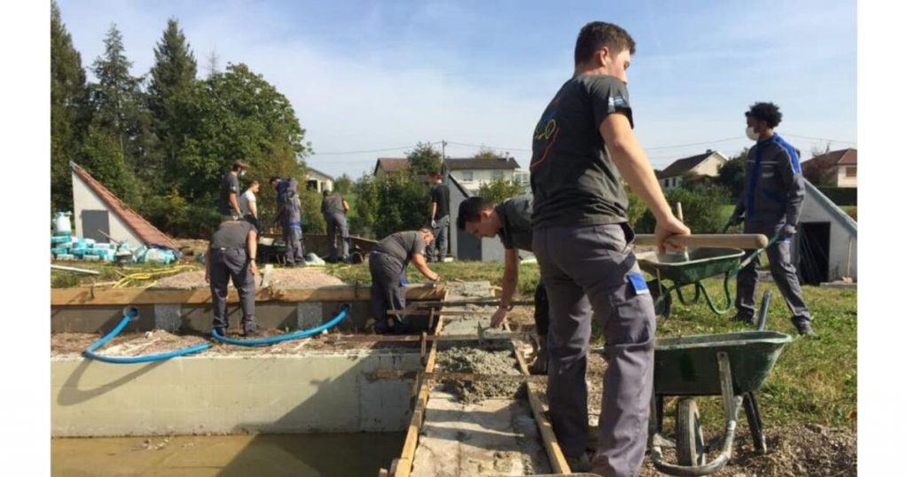 Group of apprentices and young pool technicians in uniform working on the concrete structure of a swimming pool during hands-on training in France or Belgium, illustrating apprenticeship and skill transmission in the pool industry
