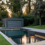 Luxury French modular swimming pool with matte anthracite-grey panels, integrated underwater roller shutter, gently sloping immersed sand-coloured beach, subtle LED lighting and sleek minimalist coping, photographed at golden hour in a contemporary garden. Symbol of 2025 French pool trends – fast installation, high-end design and total adaptability.