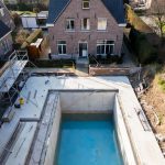 Aerial view of a high-end concrete swimming pool under construction in a typical Flemish residential backyard, Belgium. Brick gable house in background with scaffolding, materials, and blue basin ready for finishing.