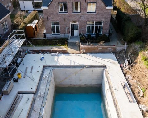 Aerial view of a high-end concrete swimming pool under construction in a typical Flemish residential backyard, Belgium. Brick gable house in background with scaffolding, materials, and blue basin ready for finishing.