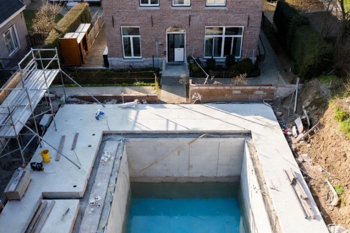 Aerial view of a high-end concrete swimming pool under construction in a typical Flemish residential backyard, Belgium. Brick gable house in background with scaffolding, materials, and blue basin ready for finishing.