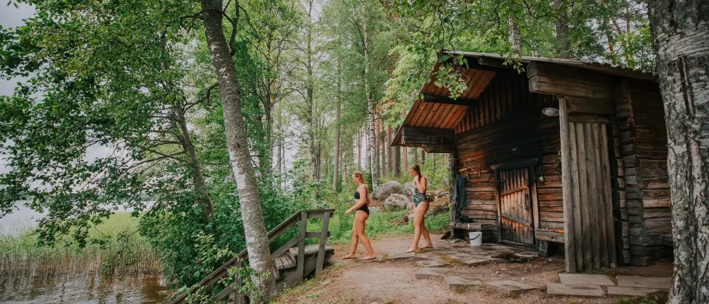Two women walking toward a traditional wooden lakeside sauna in the Finnish lake region during World Sauna Forum.