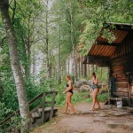 Two women walking toward a traditional wooden lakeside sauna in the Finnish lake region during World Sauna Forum.