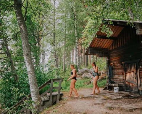 Two women walking toward a traditional wooden lakeside sauna in the Finnish lake region during World Sauna Forum.