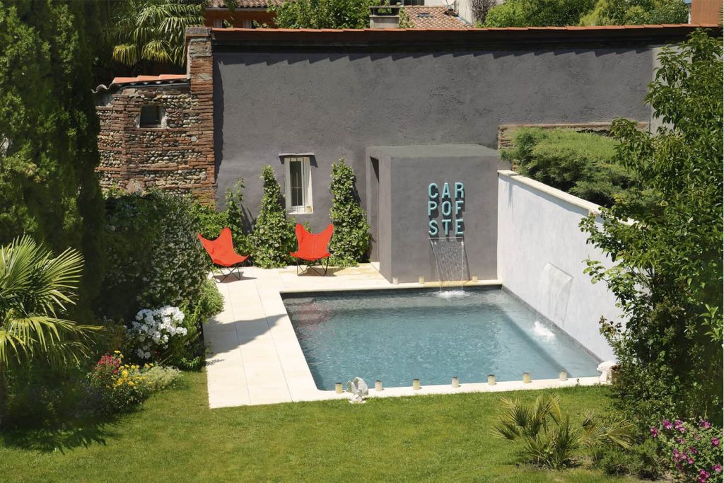 Modern small urban concrete swimming pool with two waterfall features, orange butterfly chairs, and a grey wall with "CARPE DIEM" lettering in a lush backyard garden.