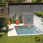 Modern small urban concrete swimming pool with two waterfall features, orange butterfly chairs, and a grey wall with "CARPE DIEM" lettering in a lush backyard garden.