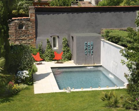 Modern small urban concrete swimming pool with two waterfall features, orange butterfly chairs, and a grey wall with "CARPE DIEM" lettering in a lush backyard garden.
