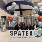 Four female members of the SPATEX organizing team standing behind a large "SPATEX 30 Years Anniversary Edition" sign in a brightly lit convention center lobby, flanked by colorful balloon displays.