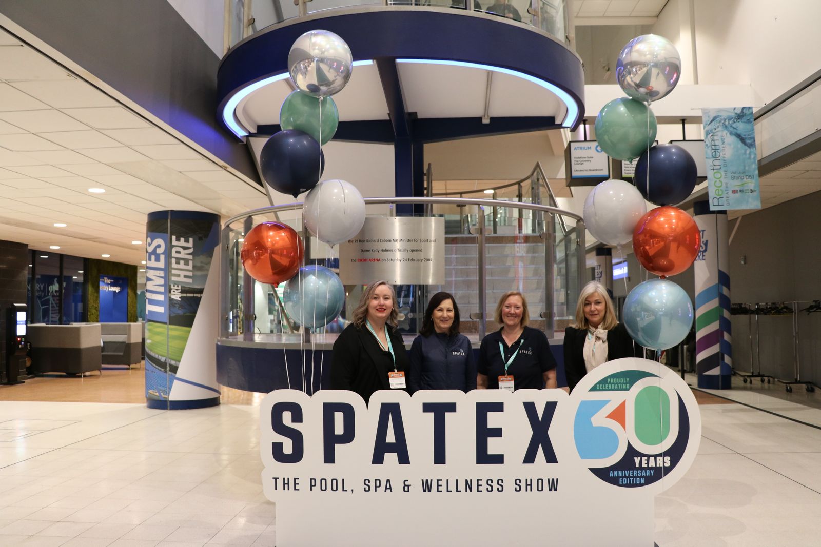 Four female members of the SPATEX organizing team standing behind a large "SPATEX 30 Years Anniversary Edition" sign in a brightly lit convention center lobby, flanked by colorful balloon displays.