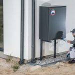 Professional technician installing a modern dark grey French-branded heat pump on a residential wall with a swimming pool background.