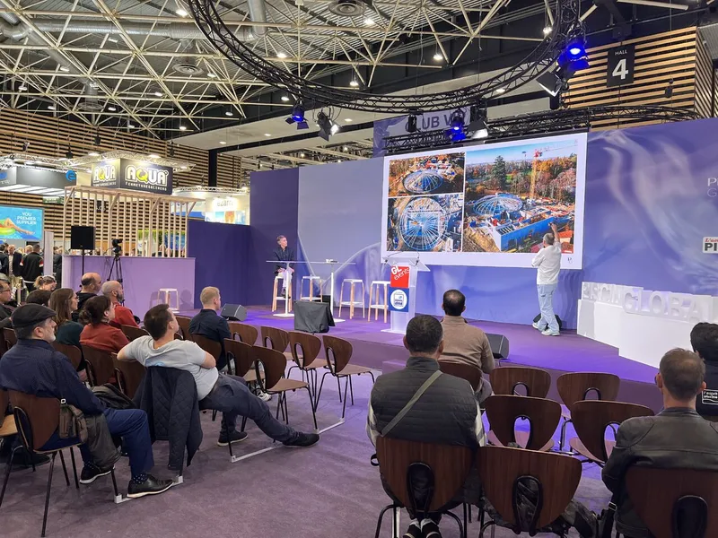 Weitblick in die Messehalle der Piscine Global auf der Eurexpo Lyon mit einem Konferenz- oder Podiumsdiskussionsbereich, in dem sitzende Fachleute, große Bildschirme und Hintergrund-Ausstellungsstände voller Pool- und Spa-Aussteller zu sehen sind, die die dynamische Networking- und Wissensaustausch-Atmosphäre der internationalen Pool-, Spa- und Wellness-Veranstaltung einfangen.