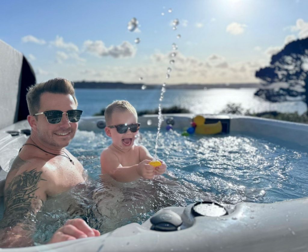 A father and son laughing together in an outdoor hot tub overlooking the sea.
