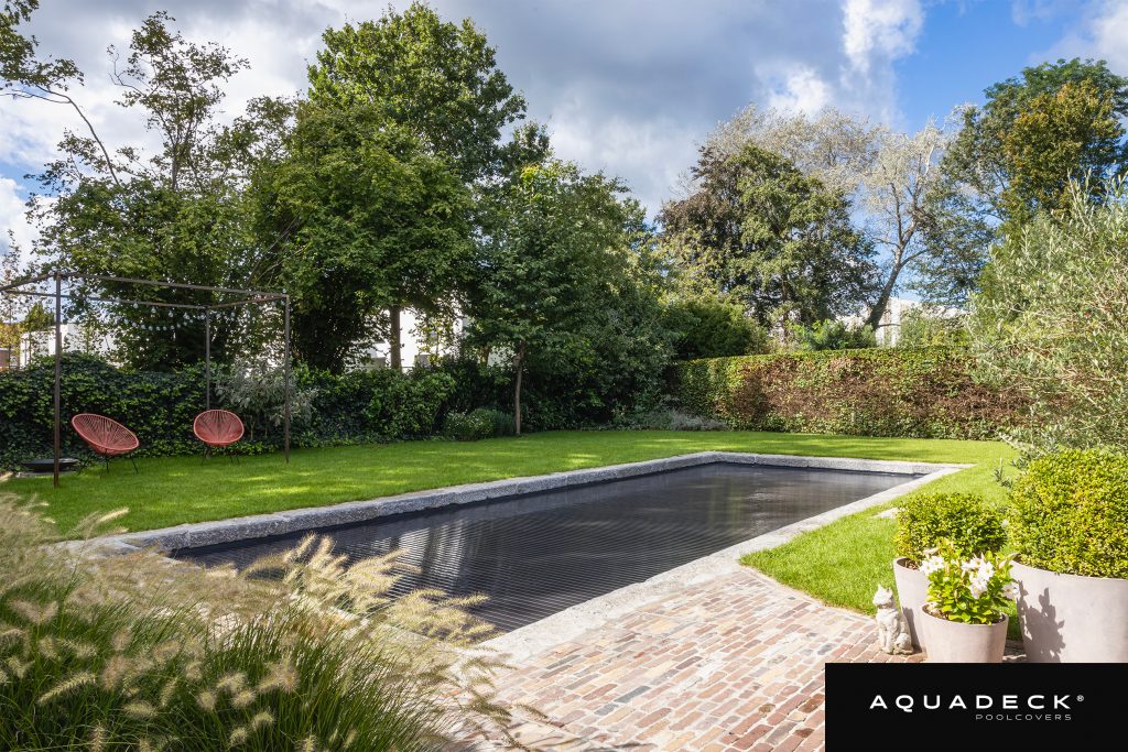 Ein moderner rechteckiger Swimmingpool mit einer dunklen Aquadeck-Automatikabdeckung aus Lamellen, installiert in einem üppigen grünen Garten mit Ziegelpflaster.