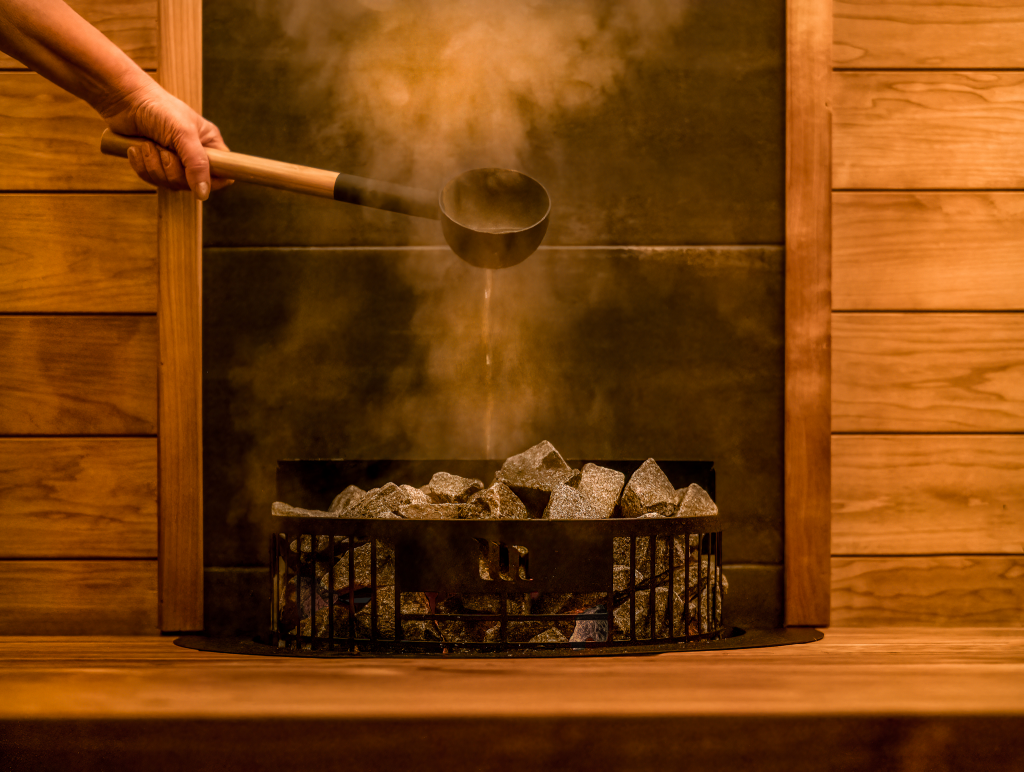 Close-up of water being poured from a wooden ladle onto hot sauna stones, creating steam in a traditional wooden sauna.