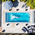 A high-angle aerial view of a person swimming in a modern, rectangular pool on a tiled patio.