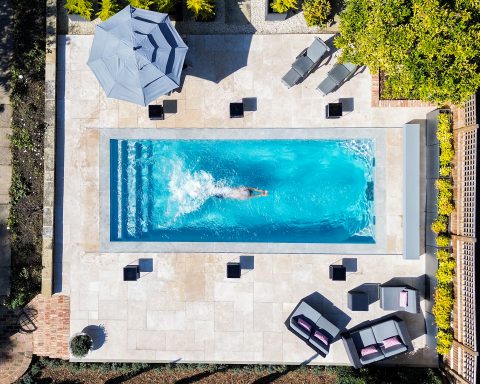 A high-angle aerial view of a person swimming in a modern, rectangular pool on a tiled patio.