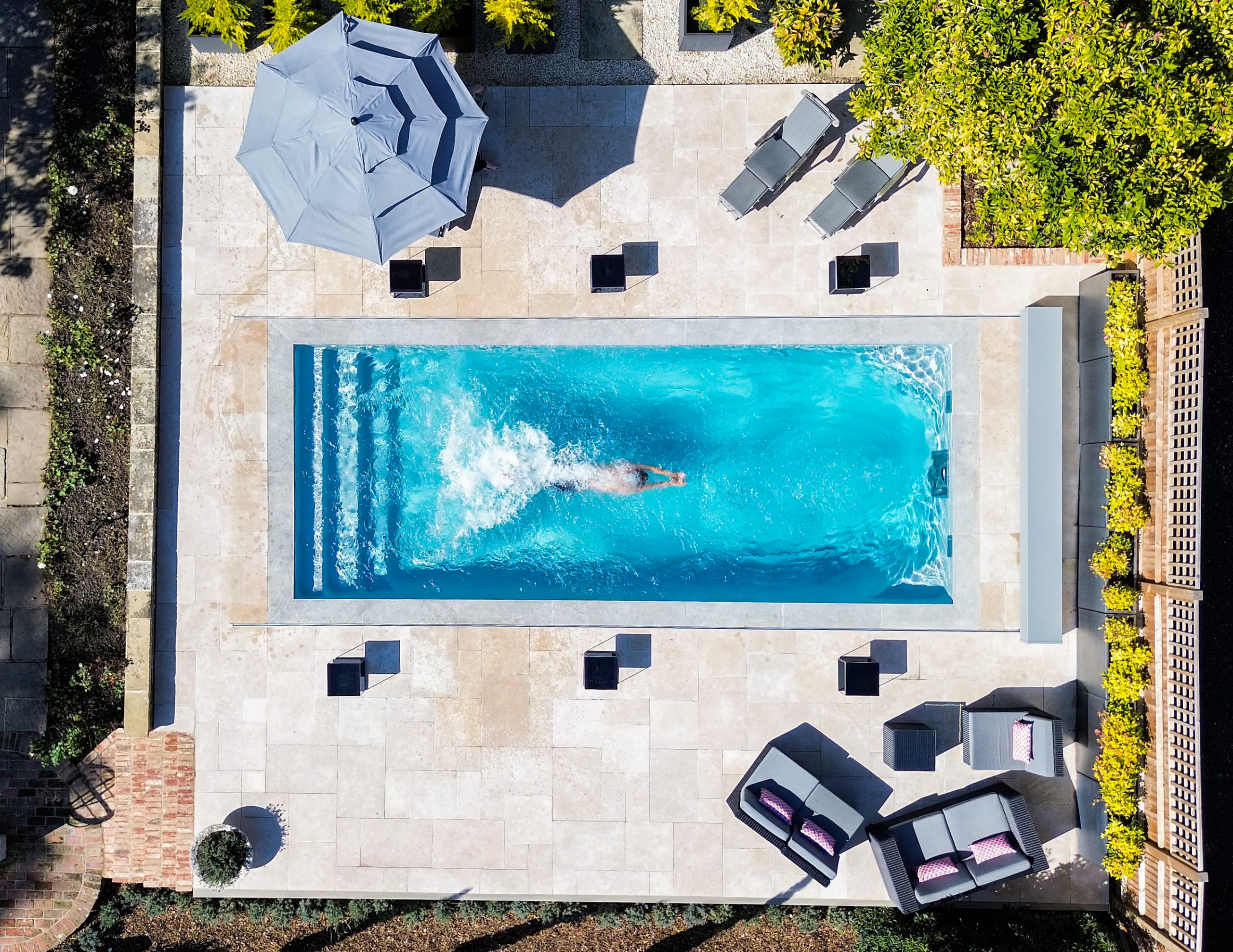 A high-angle aerial view of a person swimming in a modern, rectangular pool on a tiled patio.