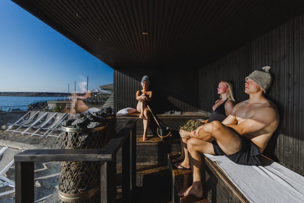 Three people relaxing in a modern, dark-wood AITO sauna at Löyly Helsinki, Finland, with a large glass window overlooking the Baltic Sea on a sunny day. A man is wearing a sauna hat, a woman is holding a birch whisk, and a distinctive pillar sauna heater with large stones is in the foreground.