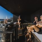 Three people relaxing in a modern, dark-wood AITO sauna at Löyly Helsinki, Finland, with a large glass window overlooking the Baltic Sea on a sunny day. A man is wearing a sauna hat, a woman is holding a birch whisk, and a distinctive pillar sauna heater with large stones is in the foreground.