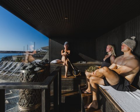 Three people relaxing in a modern, dark-wood AITO sauna at Löyly Helsinki, Finland, with a large glass window overlooking the Baltic Sea on a sunny day. A man is wearing a sauna hat, a woman is holding a birch whisk, and a distinctive pillar sauna heater with large stones is in the foreground.