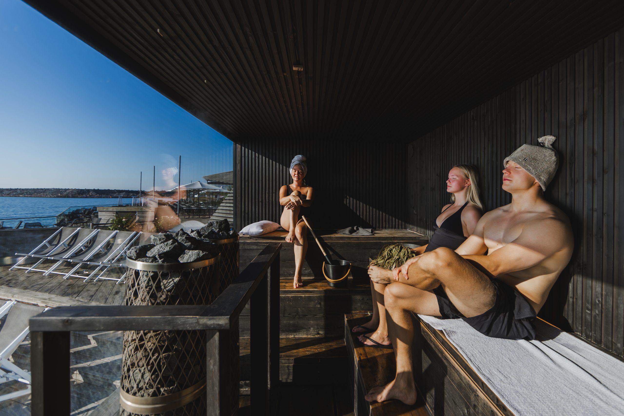 Three people relaxing in a modern, dark-wood AITO sauna at Löyly Helsinki, Finland, with a large glass window overlooking the Baltic Sea on a sunny day. A man is wearing a sauna hat, a woman is holding a birch whisk, and a distinctive pillar sauna heater with large stones is in the foreground.