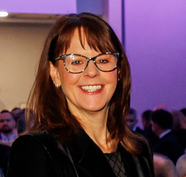 A professional portrait of Sallie Leslie-Golding wearing glasses and a black jacket, smiling in a well-lit indoor setting