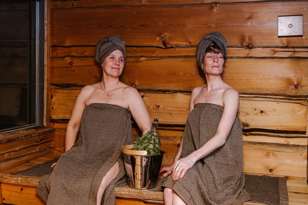 Two women relaxing in a traditional wooden sauna wearing Rento brand brown linen sauna wraps and matching head towels. They are sitting on wooden benches with a stainless steel sauna bucket and a fresh birch whisk (vihta) between them.