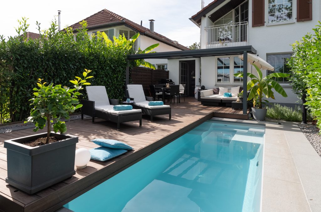 A long, narrow modern pool with a large, expansive wooden WaluDeck mobile deck and lounge furniture, set in a meticulously designed garden.