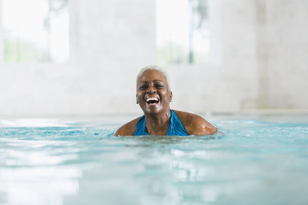A senior woman with short grey hair laughing joyfully while swimming in a clear indoor pool.