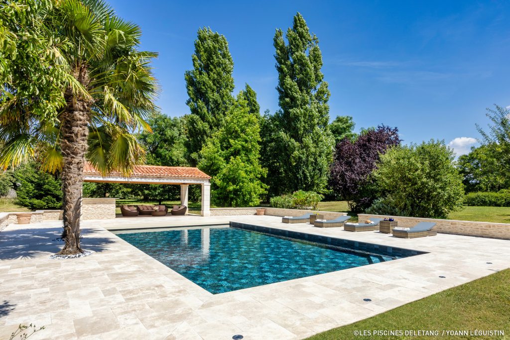 A wide view of a clean renovated rectangular swimming pool surrounded by a light-colored stone paver terrace, palm trees, sun loungers, and a pool house under a clear blue sky