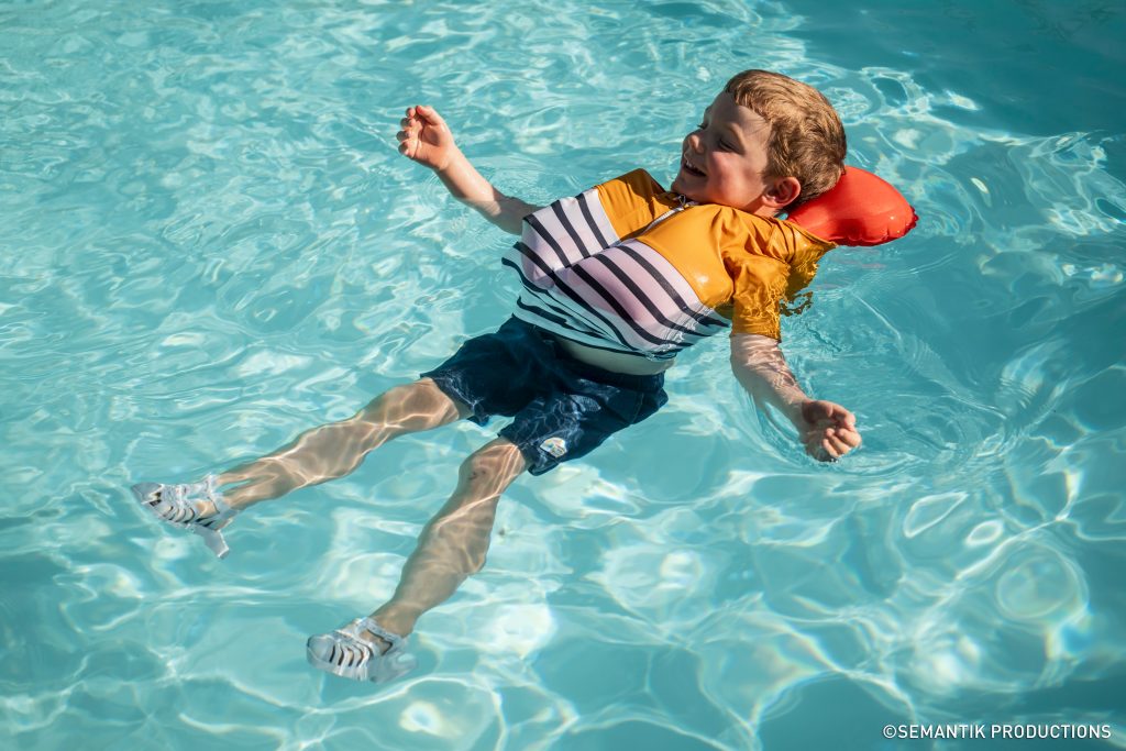A young boy smiles and floats on his back in clear blue pool water wearing a striped swim shirt and a special orange inflatable life vest