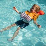 A young boy smiles and floats on his back in clear blue pool water wearing a striped swim shirt and a special orange inflatable life vest