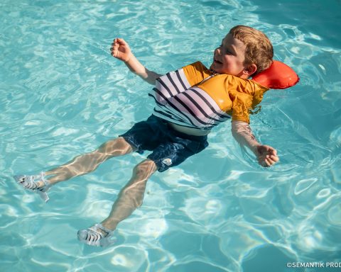 A young boy smiles and floats on his back in clear blue pool water wearing a striped swim shirt and a special orange inflatable life vest
