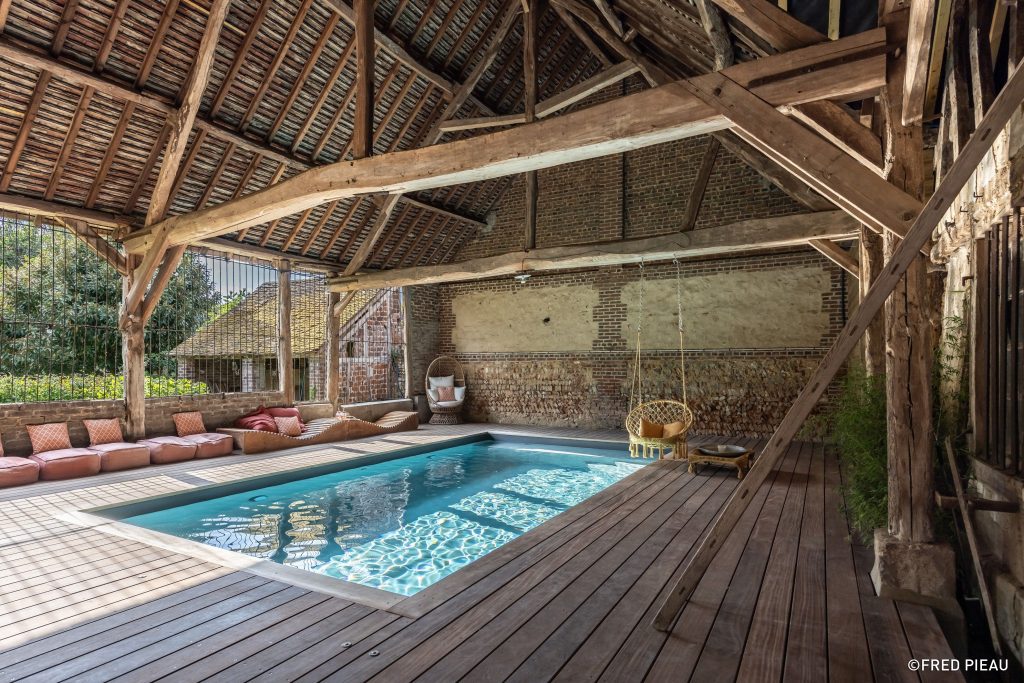 An indoor swimming pool with wooden decking inside a large rustic converted barn with massive aged timber beams, a high wooden plank ceiling, and stone walls