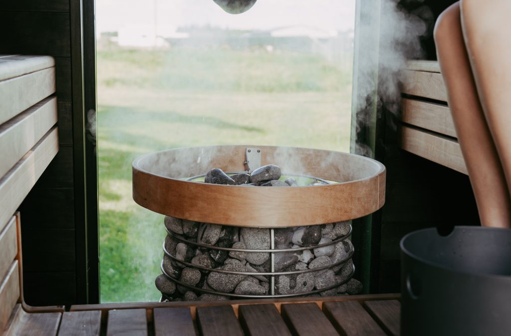 Close-up of a premium round sauna heater with volcanic stones and rising steam, glass wall overlooking a green garden