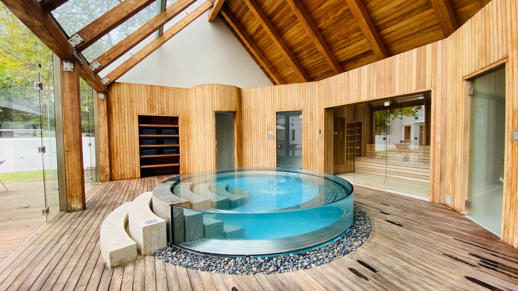Luxury indoor spa featuring a round glass-walled cold plunge pool on a wooden deck under a timber and glass roof