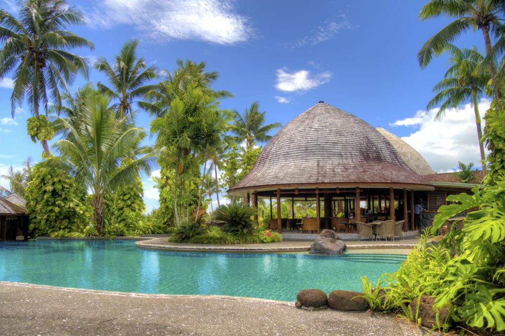 A beautiful luxury resort swimming pool surrounded by palm trees and a tropical thatched lounge area.