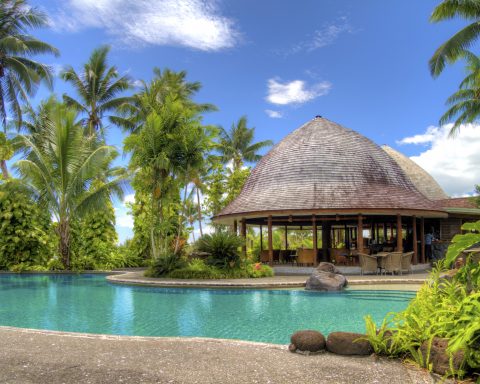 A beautiful luxury resort swimming pool surrounded by palm trees and a tropical thatched lounge area.