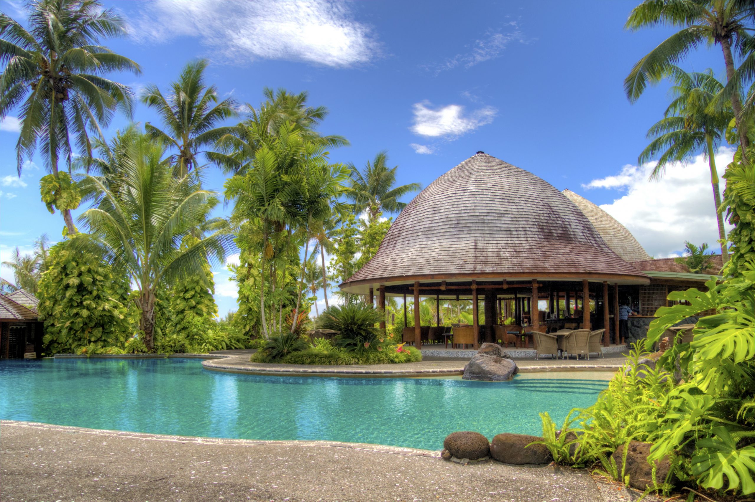 A beautiful luxury resort swimming pool surrounded by palm trees and a tropical thatched lounge area.
