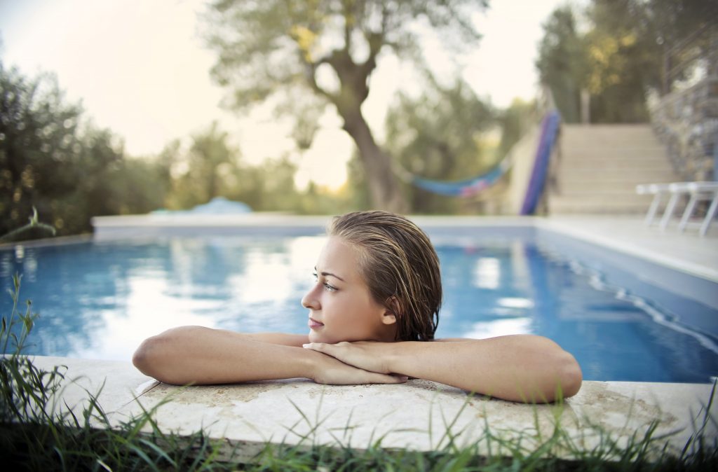 A young woman resting her arms on the stone edge of a swimming pool with a green garden in the background