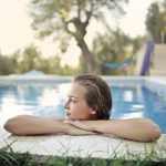 A young woman resting her arms on the stone edge of a swimming pool with a green garden in the background