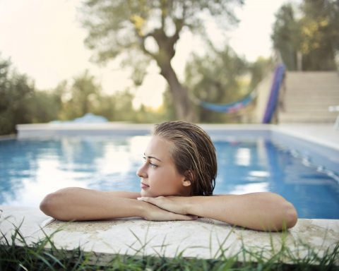 A young woman resting her arms on the stone edge of a swimming pool with a green garden in the background
