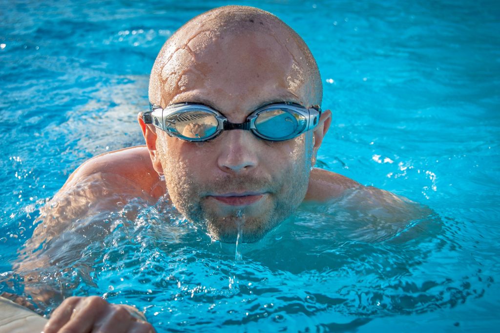 Close up portrait of a male swimmer wearing goggles coming out of a vibrant blue pool