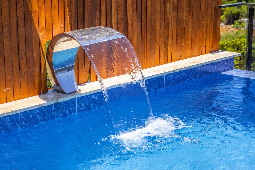 Close-up of a curved stainless steel waterfall feature pouring water into a blue tiled swimming pool against a wooden fence.
