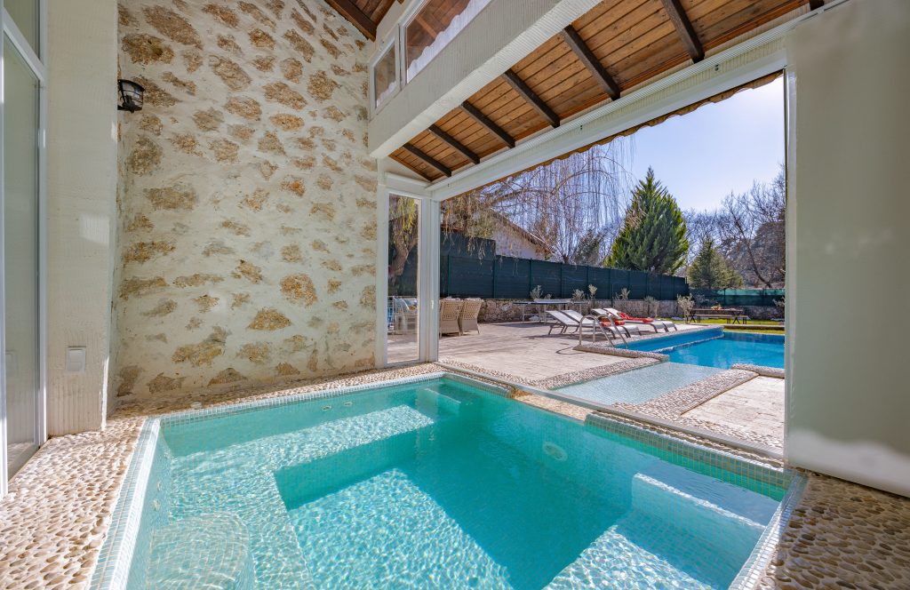 Luxury indoor hydrotherapy spa pool made of stone, opening out to a sunny backyard pool deck.