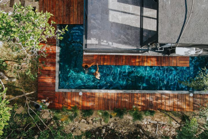 Aerial drone shot of a lap pool surrounded by modern wooden decking and lush green tropical foliage.
